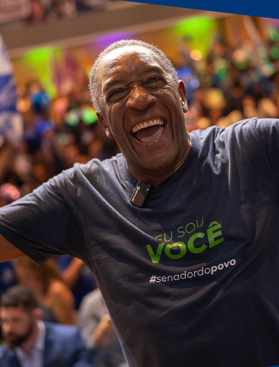 Geraldo Rufino de braços abertos, sorrindo, diante de uma plateia, usando camiseta com a frase Eu sou você.